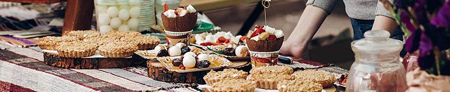 A table with various baked goods, including tarts and sweets, displayed on rustic wooden slabs. The background shows a person’s arm reaching across the table, and there are jars and other decorative items adding to the scene.