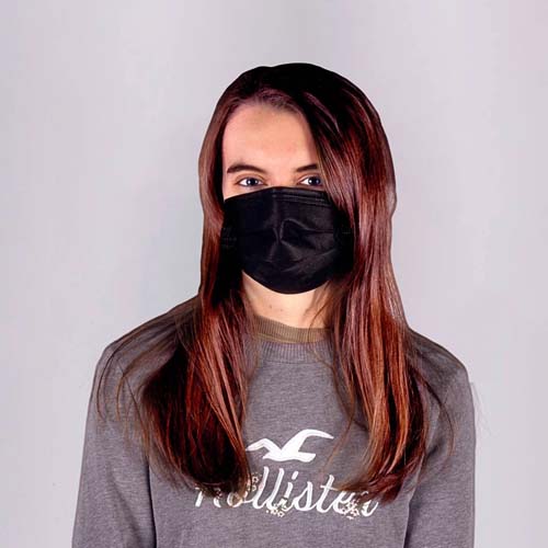 A woman with long brown hair wearing a black Type IIR medical face mask, looking directly at the camera. She is dressed in a grey sweatshirt with a logo, set against a plain, light-coloured background.