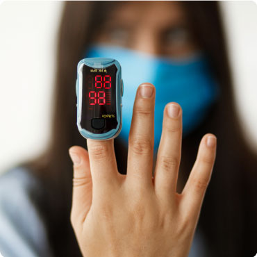 Person with C19 Finger Tip Pulse oXimeter on their finger. Their hand is in front of their face, their face is blurred out with the hand in focus