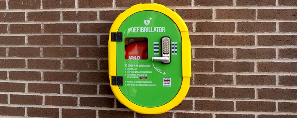 A green and yellow defibrillator case mounted on a brick wall. The case has a keypad lock and includes emergency instructions for accessing the device.
