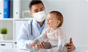Doctor with baby in exam room. The baby is laughing and doctor is smiling with a face mask on