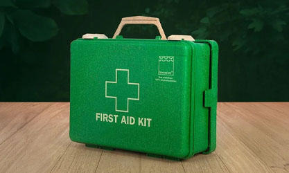 A green eco-friendly first aid kit with a handle, displayed on a wooden surface with a dark green leafy background. The kit features a white cross and the Steroplast logo.