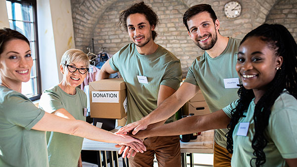 A group of diverse volunteers smiling and placing their hands together in a stack. They are dressed casually with donation boxes visible in the background, suggesting a team effort in a fundraising or charity setting.