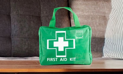 A green fabric first aid kit with a handle and a white cross, placed on a wooden table in a cosy home setting with a sofa and cushions in the background.