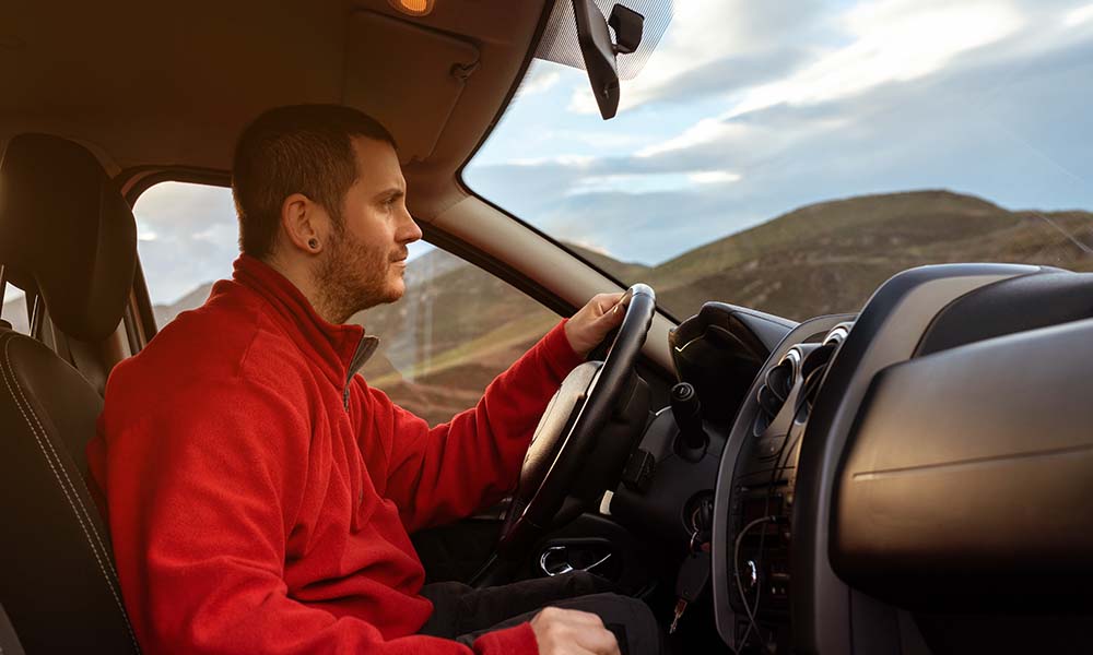A man wearing a red jacket driving a car through a scenic landscape, with green hills visible through the windows.