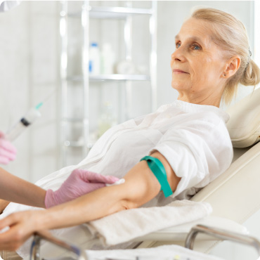 Smiling relaxed elderly female patient sitting on couch with tourniquet. Professional medical worker administering intravenous injection