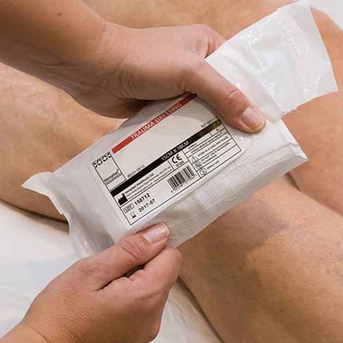 Close-up of hands holding a packaged trauma dressing, preparing to apply it. The package label is visible, and the person's legs are in the background, suggesting a medical or first-aid setting.