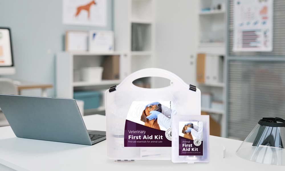 A veterinary first aid kit displayed on a desk in a veterinary office, alongside a laptop and protective equipment. The kit has a white case with a purple label showing a gloved hand examining a dog.