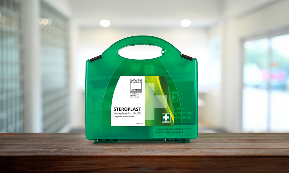 A green workplace first aid kit with a handle, labelled 'Steroplast Workplace First Aid Kit' and compliant with BS8599-1. The kit is displayed on a wooden surface with a bright office background.