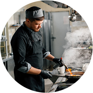 Male chef in a kitchen preparing food, with steam rising from the pan