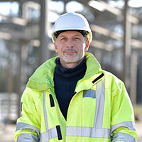 Mature man in a hard hat and high-visibility jacket, standing outdoors at a construction site