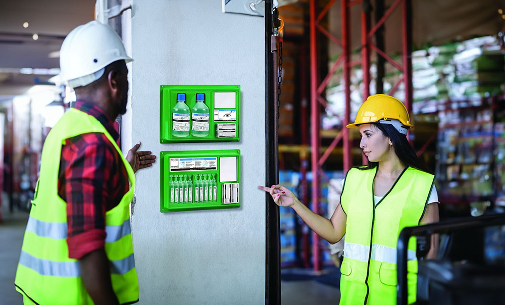 Two workers in high-visibility vests and helmets standing near an eye care station mounted on a wall in a warehouse