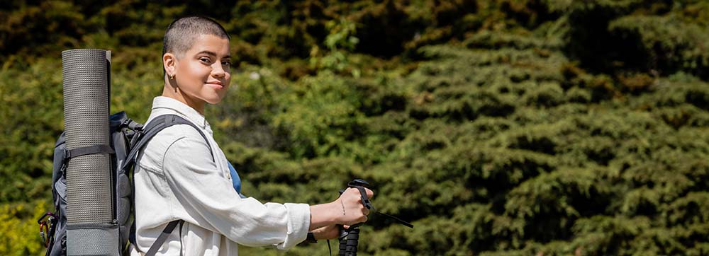 A person with short hair carrying a backpack and holding hiking poles, standing outdoors with lush green foliage in the background, suggesting a scenic hiking or nature experience.