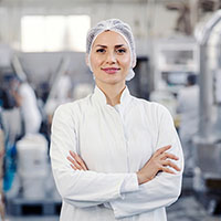 Woman in a white lab coat and hairnet, standing confidently with arms crossed in an industrial production facility