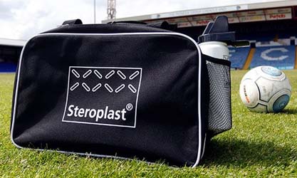 A black Steroplast sports bag featuring the Steroplast logo, placed on a grassy football field with a water bottle and football in the background.