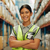 Young woman in a high-visibility vest, standing with arms crossed in a warehouse aisle, surrounded by stacked goods