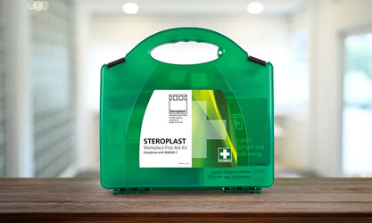 A green workplace first aid kit with a handle, labelled 'Steroplast Workplace First Aid Kit,' displayed on a wooden surface with a bright office environment in the background.