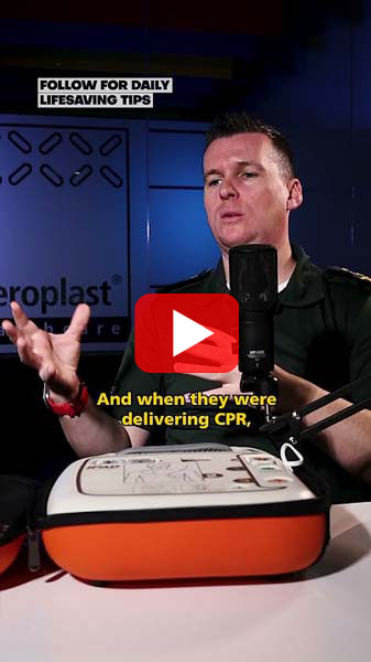 A man in a green uniform talks into a microphone with an AED device in an orange case placed in front of him. Subtitles on the screen read, 